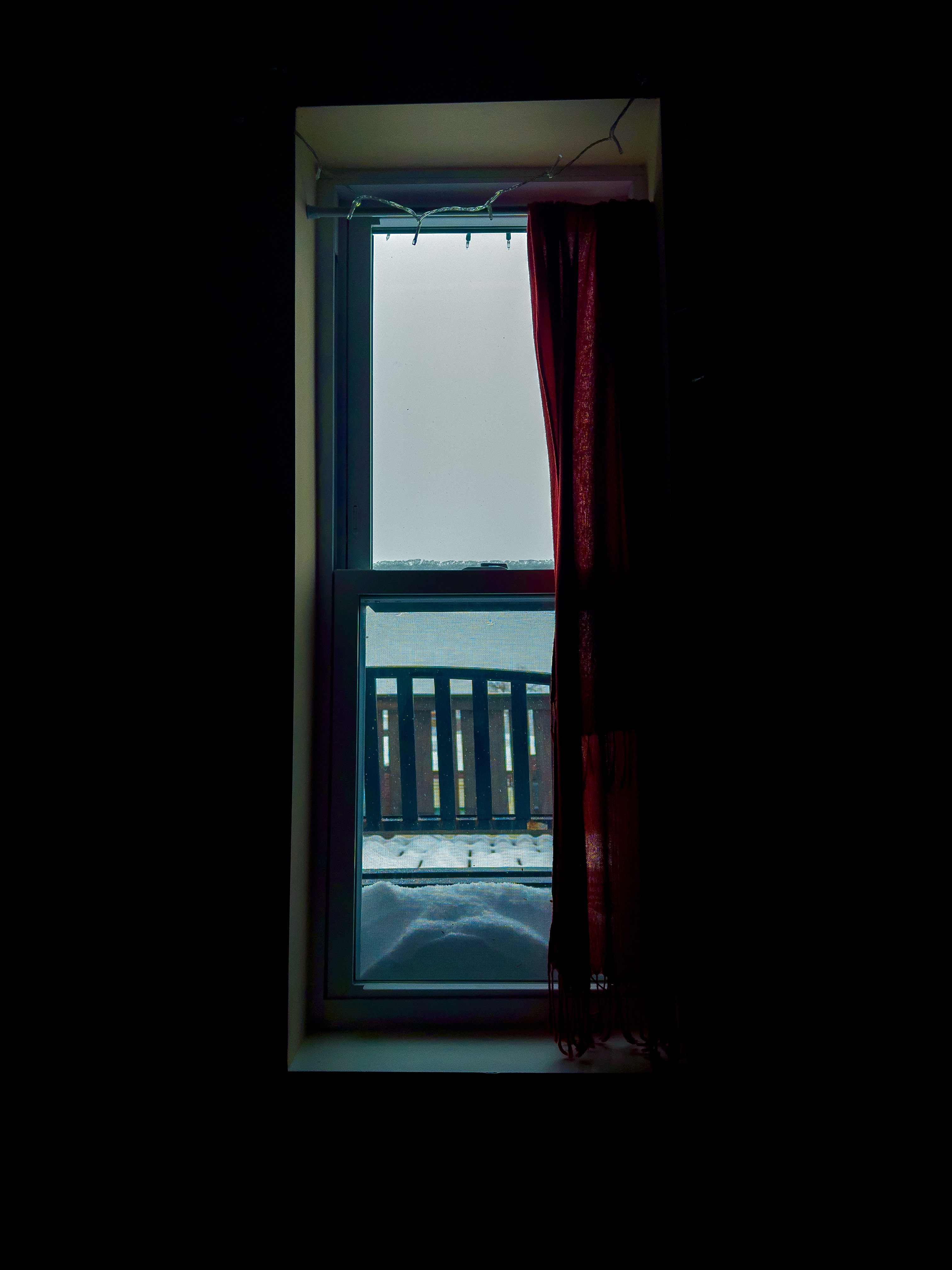 Photo of a bedroom window framed by a red curtain. The room within is dark, and looking out is a gray, dim scene of snow piling on the balcony.