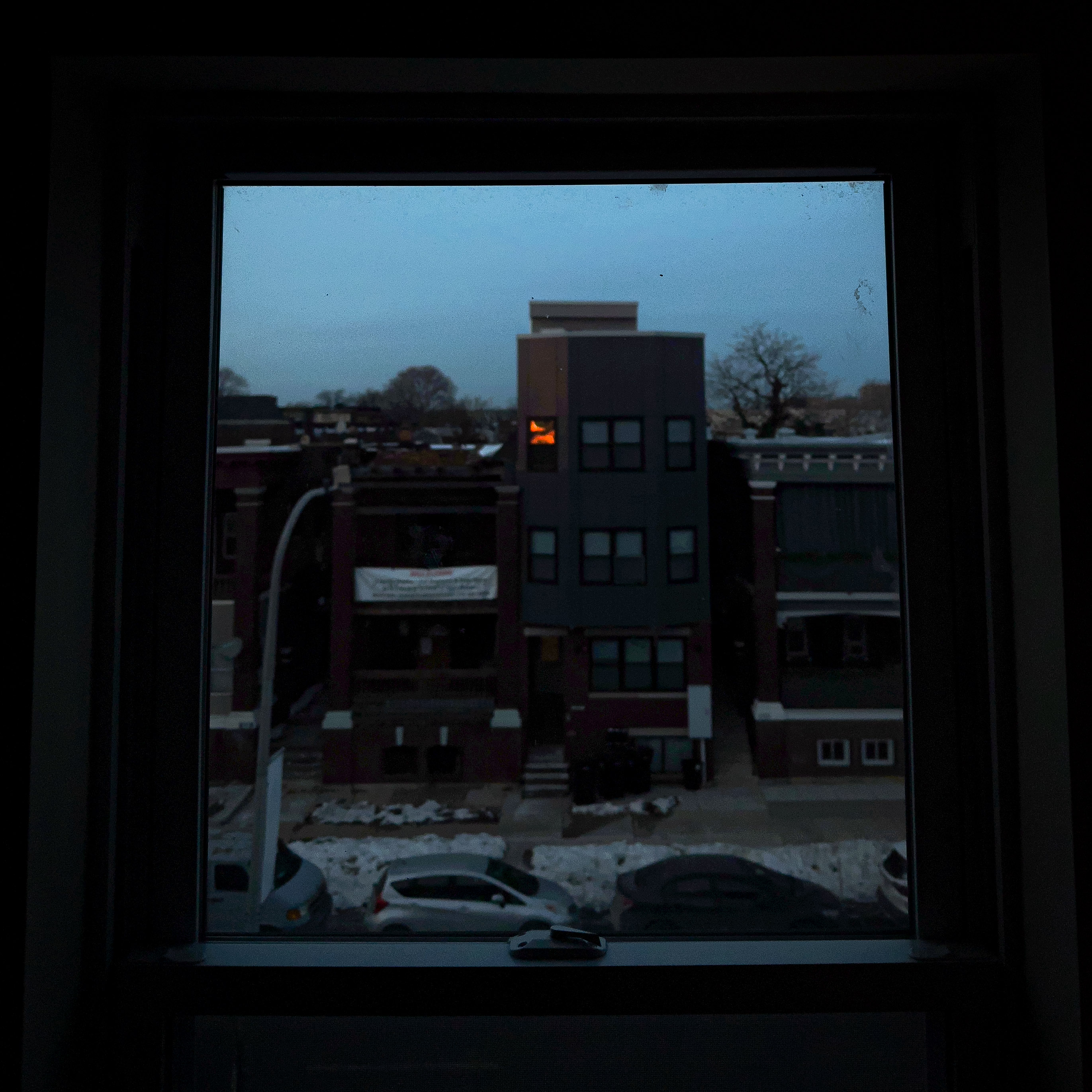 Photo of rowhomes shot through a window. The window is in focus and the buildings remain out of focus. The orange colors of the setting sun can be seen reflected in the window of a house across the street.

